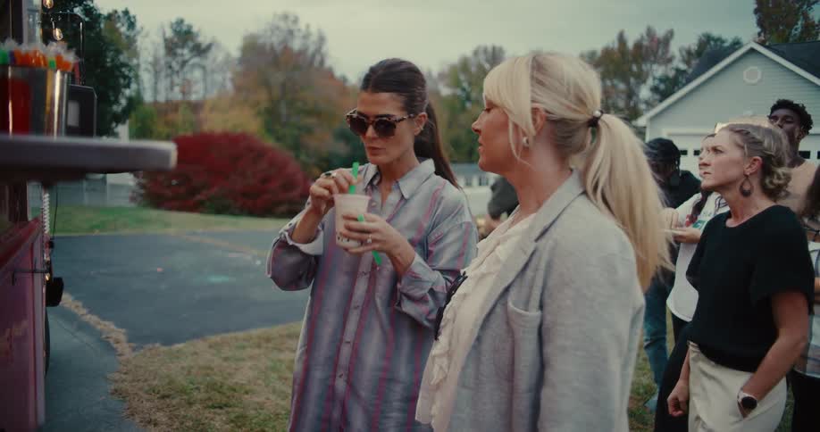 A woman in sunglasses stirs and drinks a boba tea while waiting with friends at a food truck