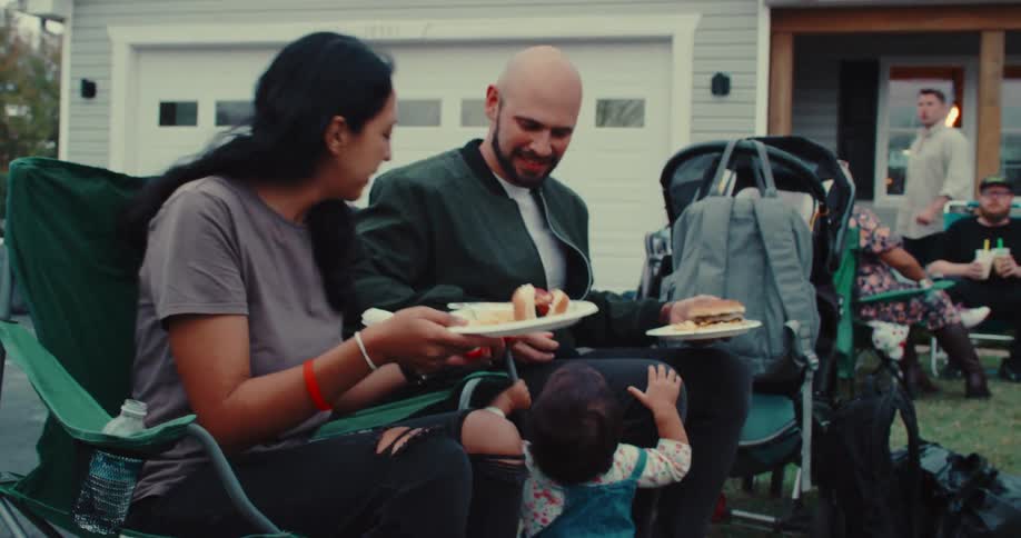 A young family with their toddler enjoys eating hot dogs and talking with friends at a neighborhood block party