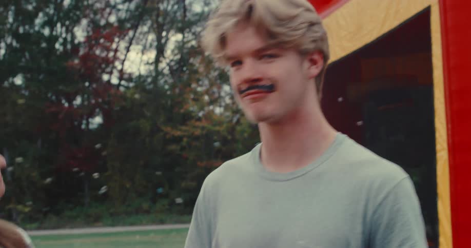 A young man gets a mustache painted on his face at a carnival, then smiles and points playfully at the camera