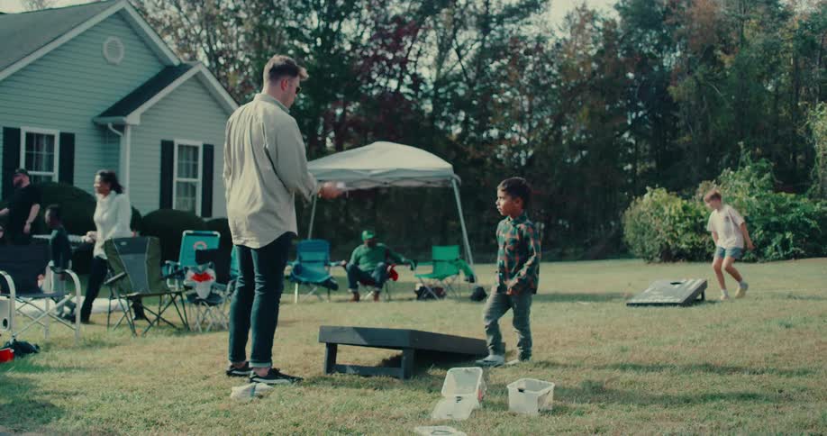A young boy and a man play a game of cornhole in the backyard during a family gathering in slow motion