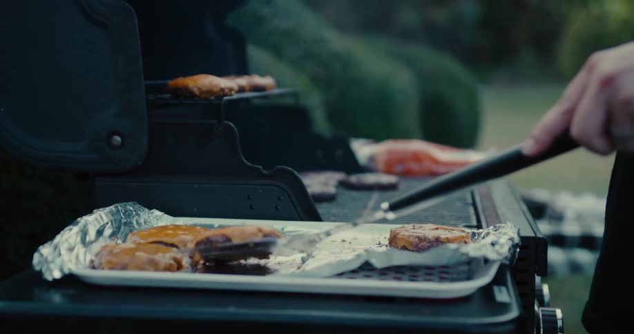 A person removes cooked cheeseburgers from the barbecue grill onto a tray with aluminum foil in slow motion