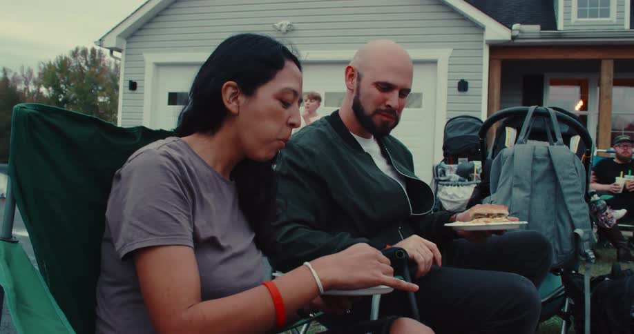 A man and a woman sit in lawn chairs and enjoy a meal at an outdoor gathering in a suburban neighborhood at dusk