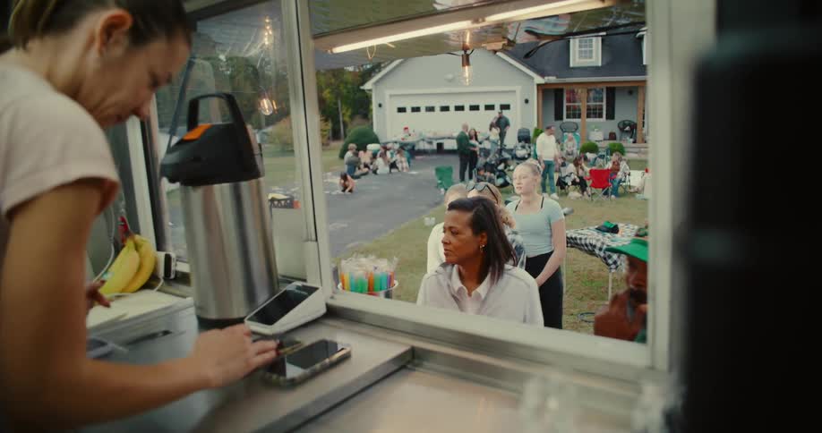 An over-the-shoulder view from inside a food truck as a vendor takes an order from a diverse group of people at a neighborhood block party