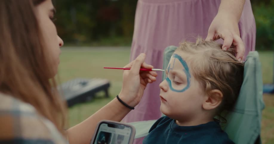 A young boy sits patiently while having a blue superhero mask painted on his face at an outdoor event