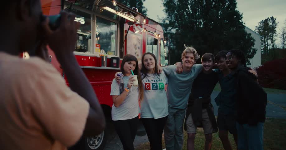 A diverse group of smiling teenage friends pose for a photo in front of a red food truck at dusk
