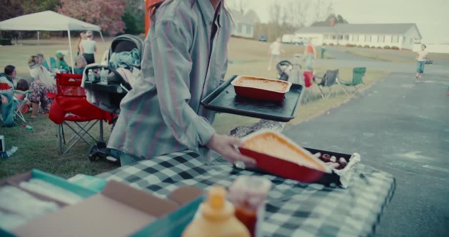 In slow motion, a woman brings a homemade casserole to a lively backyard block party with friends and family