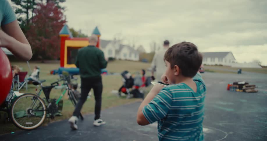 A young boy smiles while eating a hot dog at an outdoor block party with a bouncy house in the background, filmed in slow motion