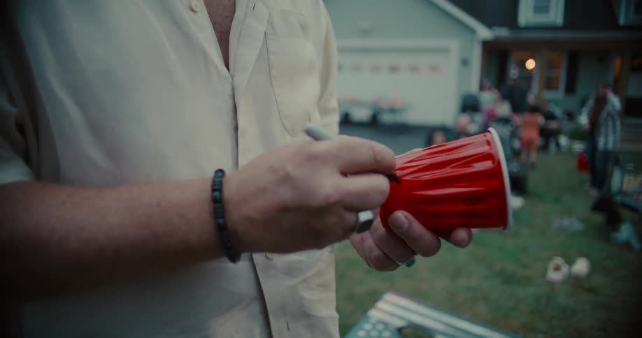 In slow motion, a man writes his name on a red plastic cup at a backyard party as people celebrate in the background