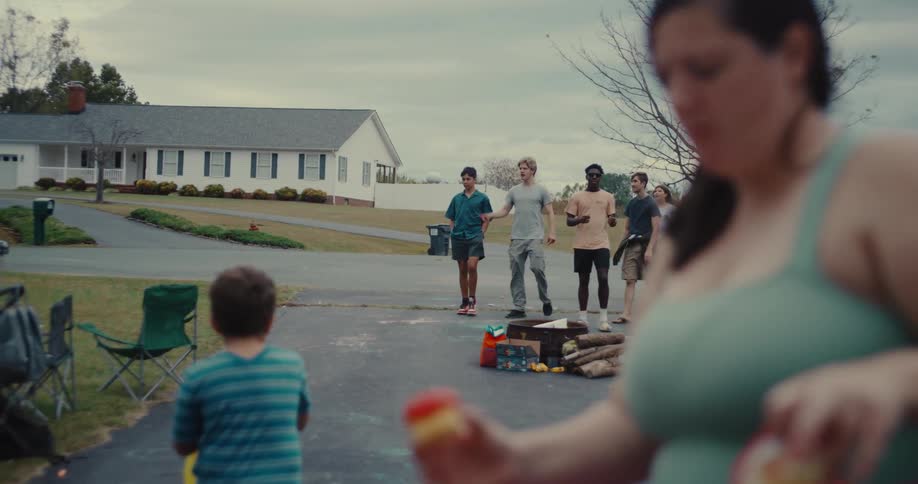 A diverse group of people gather in a suburban driveway for a neighborhood block party in slow motion