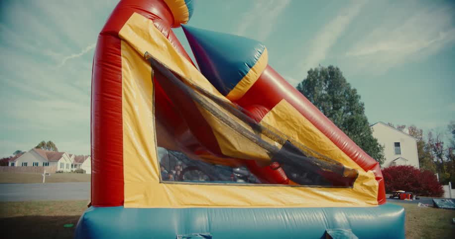 A man turns on a blower to inflate a large, colorful bounce house, which children then happily jump inside at an outdoor party