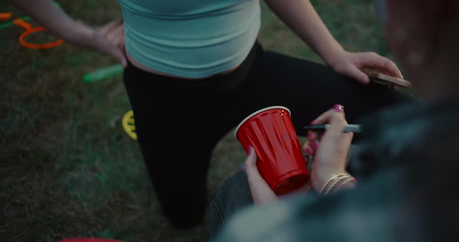 A young woman writes on her red party cup with a permanent marker while sitting on the grass