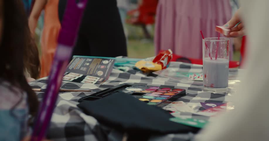 A close-up of a face painting station at an outdoor children's party with hands dipping brushes into a colorful palette