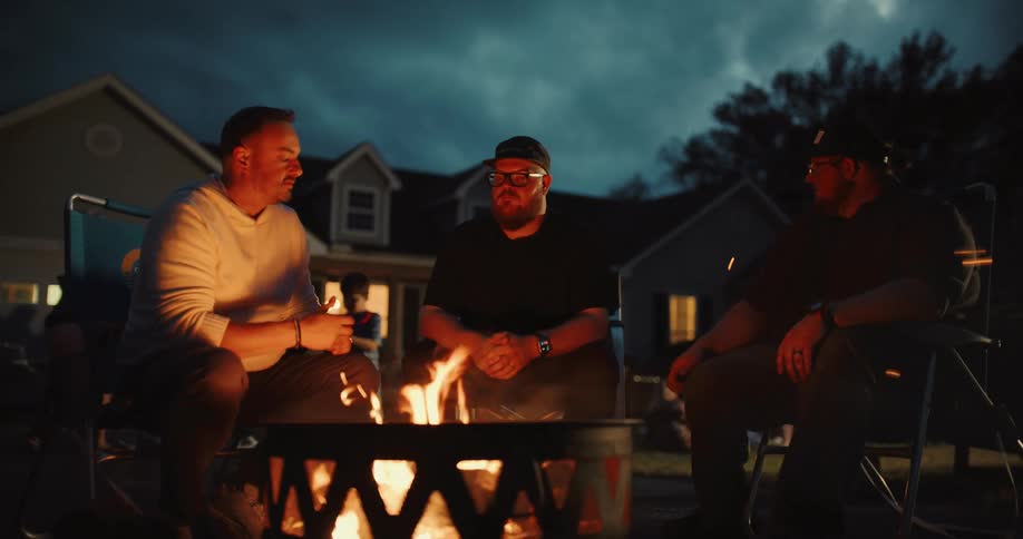 Three male friends share stories and conversation while sitting around a cozy fire pit during a backyard gathering at dusk