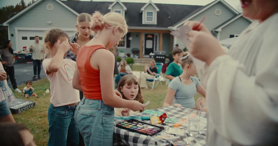 A young girl applies face paint to her friend at a fun community gathering in a suburban backyard
