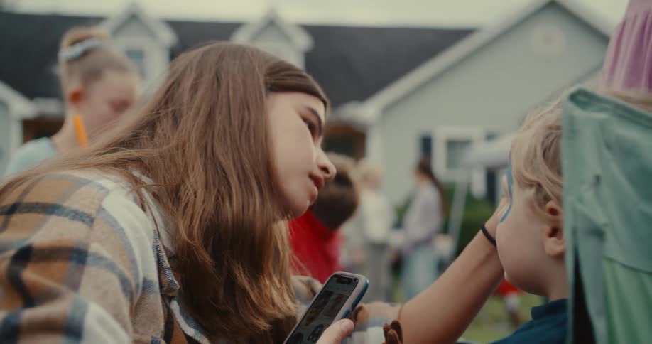 A young woman uses her smartphone for reference as she carefully applies face paint to a child at an outdoor party