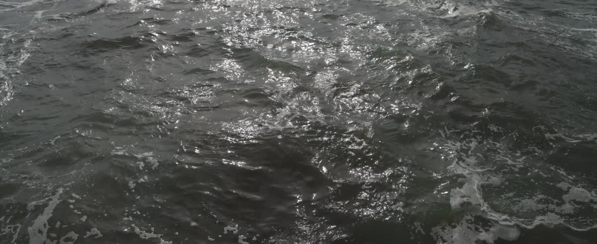 Sunlight reflects and shimmers across the dark, undulating surface of the ocean in slow motion