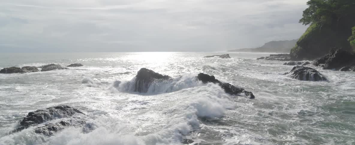 Slow motion footage of powerful ocean waves crashing over dark volcanic rocks on a bright, overcast day