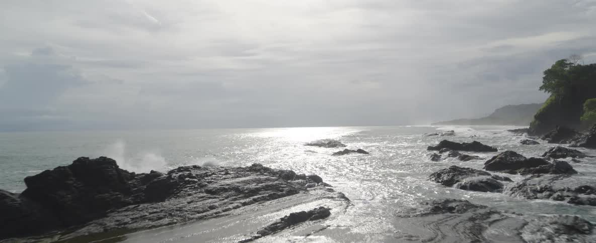 The bright sun glistens on the vast ocean as the camera pulls back to reveal a rugged, rocky coast