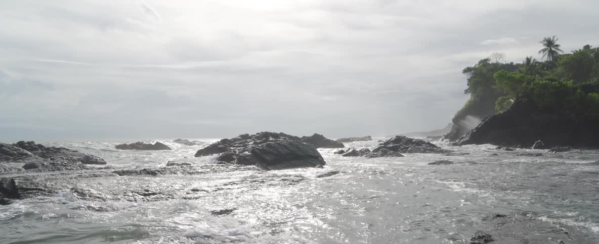 The tide slowly rises, submerging the dark, jagged rocks along a misty coastline under a bright, cloudy sky