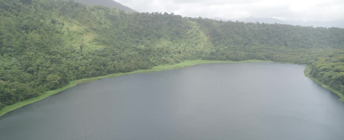 A static high-angle view of a tranquil crater lake surrounded by a lush green rainforest on a hazy, overcast day
