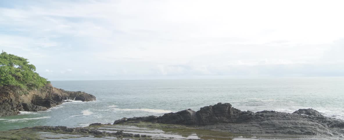 Gentle ocean waves lap against a rocky coastline under a bright, cloudy sky