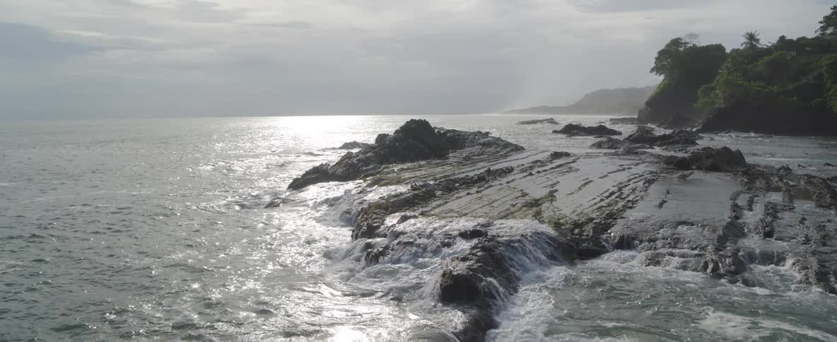 Gentle ocean waves wash over a rugged, rocky coastline as the bright sun reflects on the water's surface
