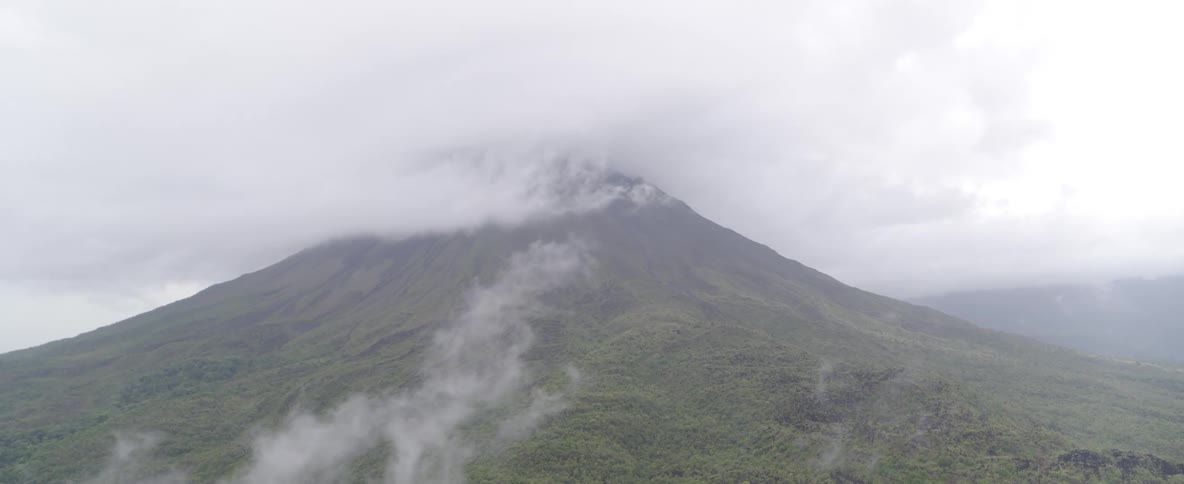 A stationary wide shot captures a large, forested volcano with its peak hidden by thick clouds and fog on an overcast day
