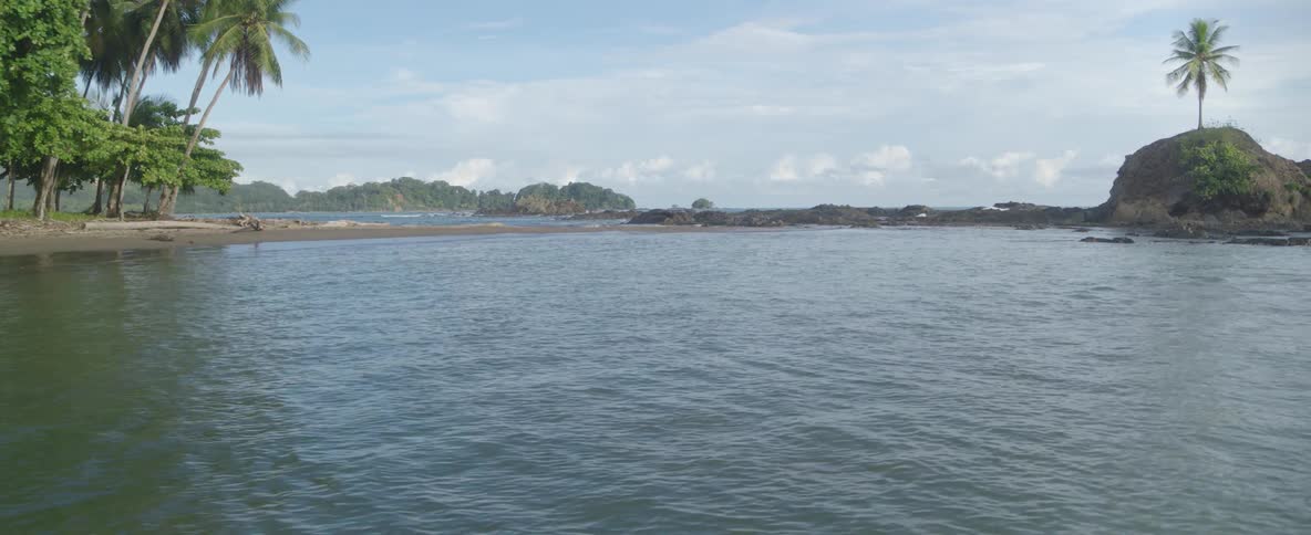 A serene view of a tropical bay with a sandy, palm-lined shore and a rocky outcrop separating it from the ocean
