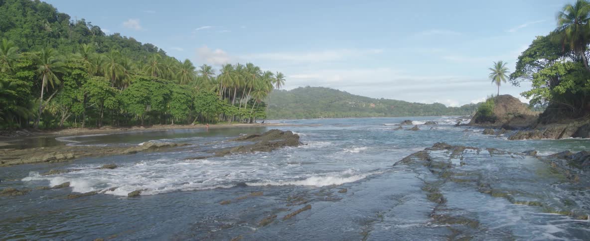 A slow aerial pullback reveals a stunning tropical bay with waves washing over unique layered rock formations at low tide