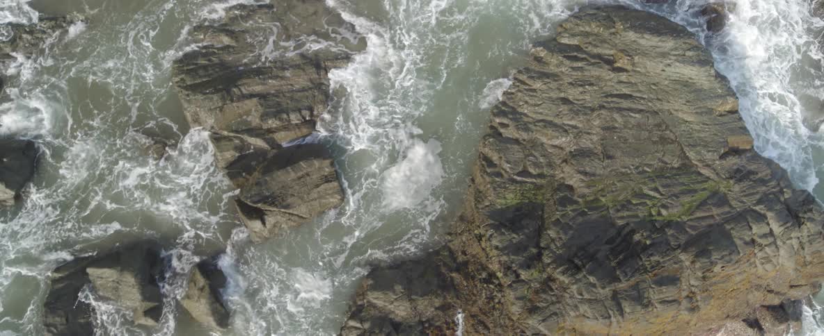 A slowly ascending overhead drone shot reveals the powerful ocean swirling and crashing into a rocky shoreline