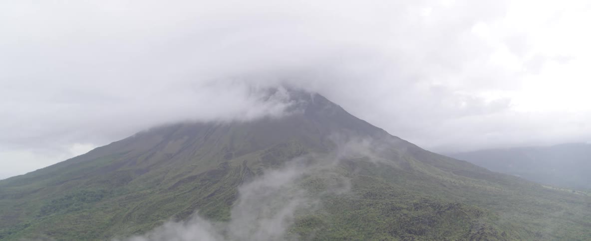 A static view captures a majestic green volcano, its peak hidden by a dense layer of rolling clouds and mist
