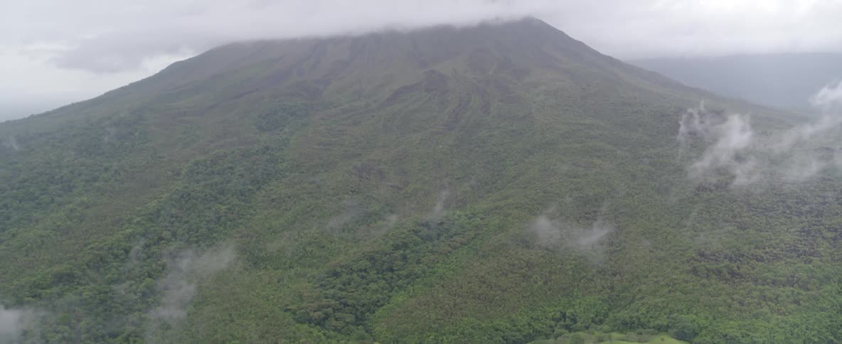 Aerial shot tilts up from a lush green jungle and a small pond to reveal a massive volcano shrouded in clouds