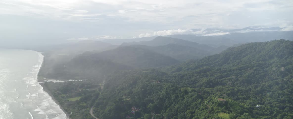 An expansive aerial shot of a misty tropical jungle cascading down to meet the ocean's shoreline