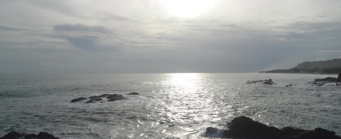 A slow aerial pullback reveals a dramatic rocky coastline with the bright sun reflecting on the ocean waves