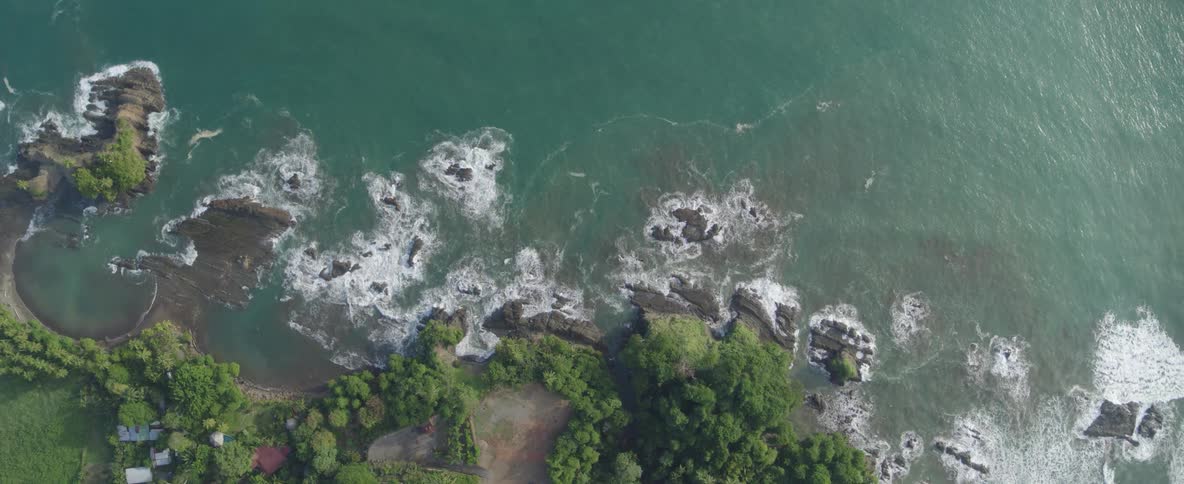 A slow ascending aerial top-down view captures waves crashing on a rocky, tropical coastline