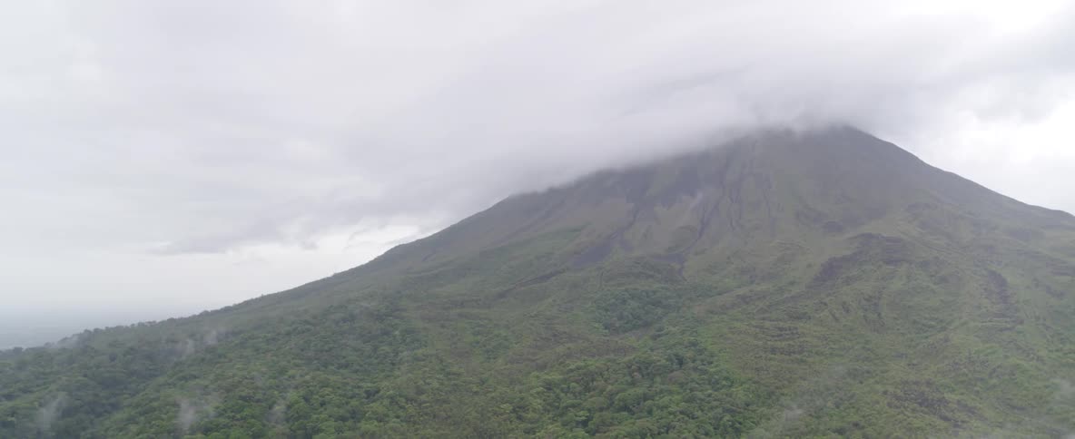 A slow aerial pan reveals a massive volcano with its peak shrouded in thick clouds above a dense, misty rainforest