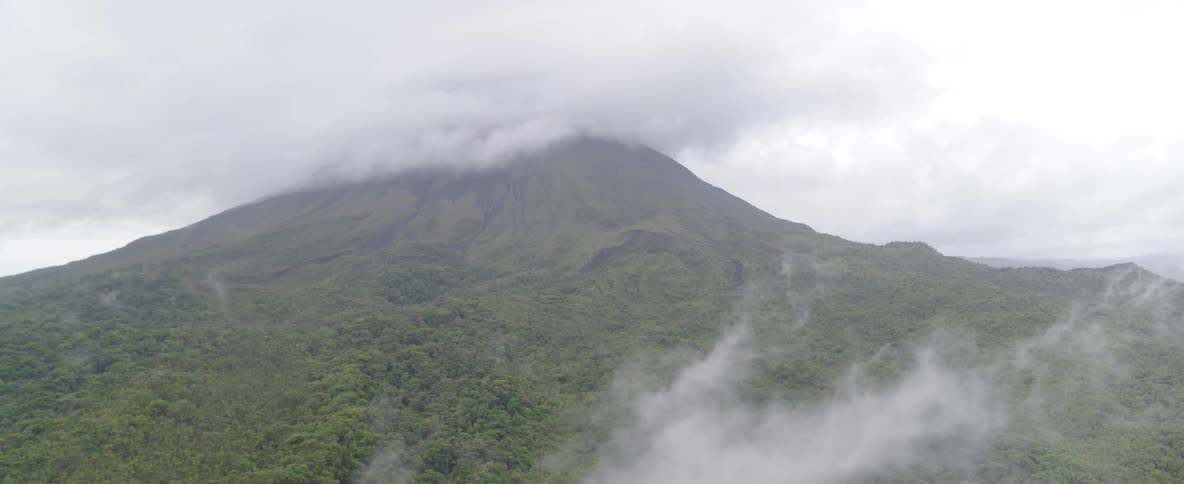 A majestic green volcano with its peak hidden in the clouds rises above a dense forest on a foggy day