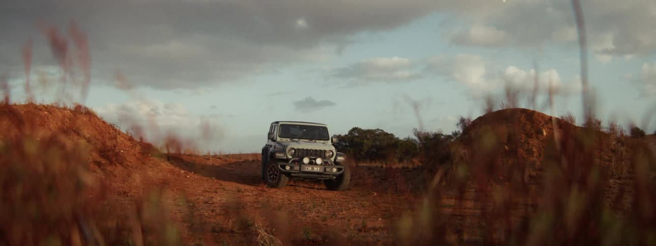 A rugged white Jeep is parked on a remote red dirt road under a vast, cloudy sky at dusk