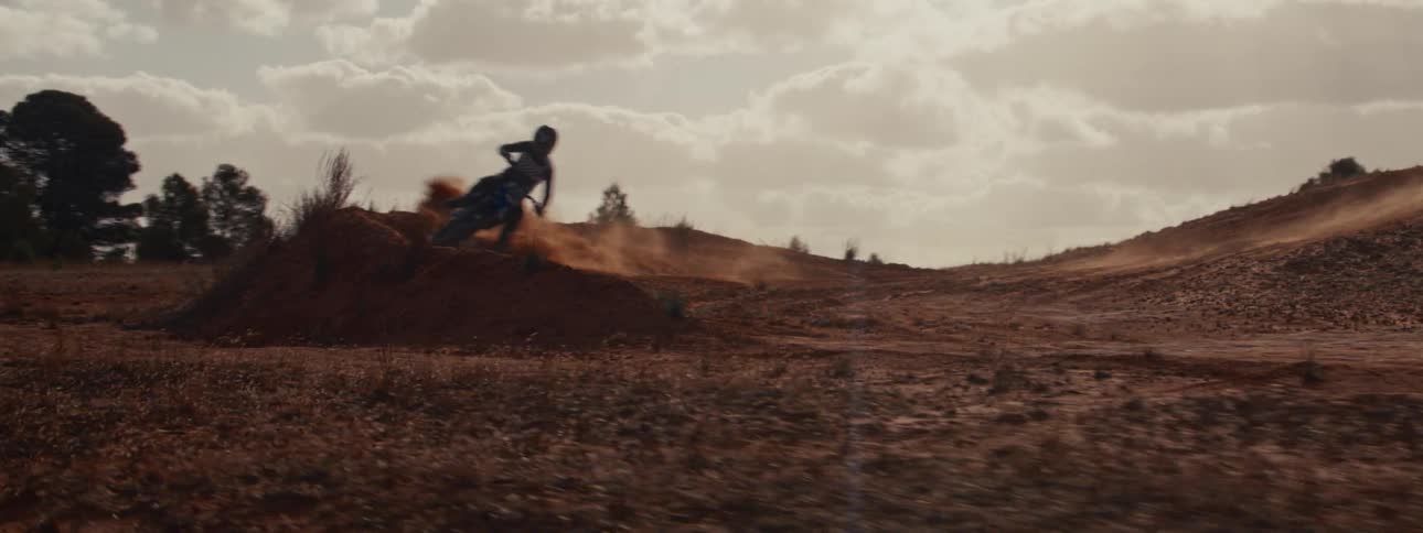 Slow motion tracking shot of a motocross rider making a sharp turn on a dirt track, kicking up a cloud of red dust