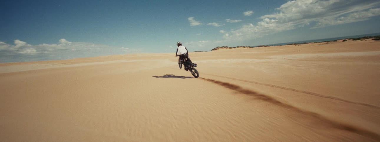 Slow motion tracking shot follows a motocross rider as they speed, jump, and wheelie across expansive sand dunes