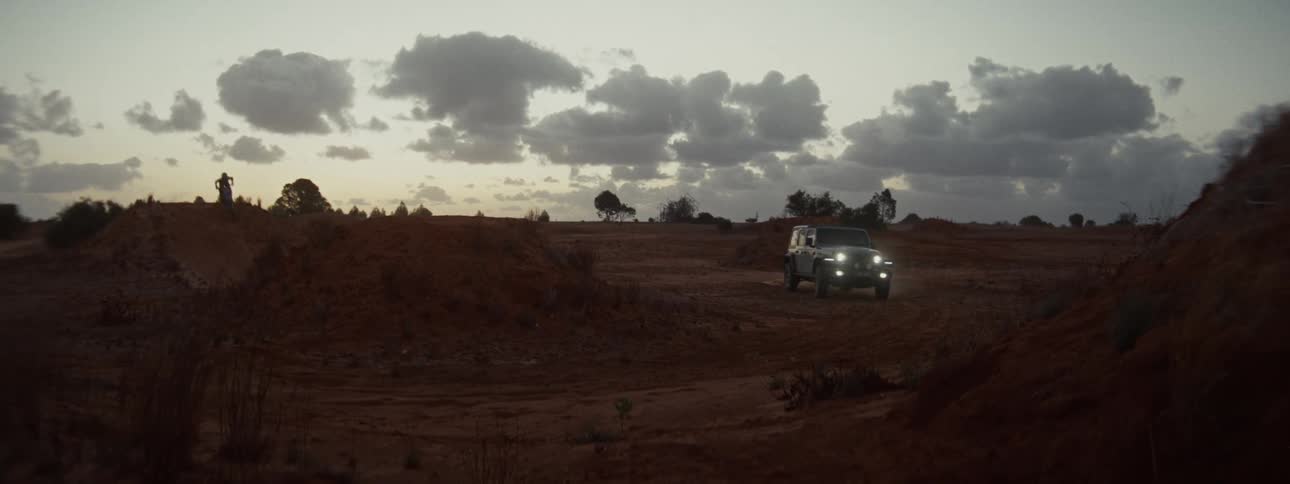 A dirt bike rider performs a dramatic jump over a dirt mound as an SUV drives through the dusty outback at dusk