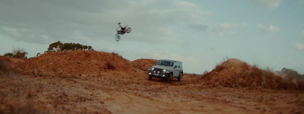A thrilling shot reveals a motocross biker jumping over a parked Jeep on a rugged dirt track