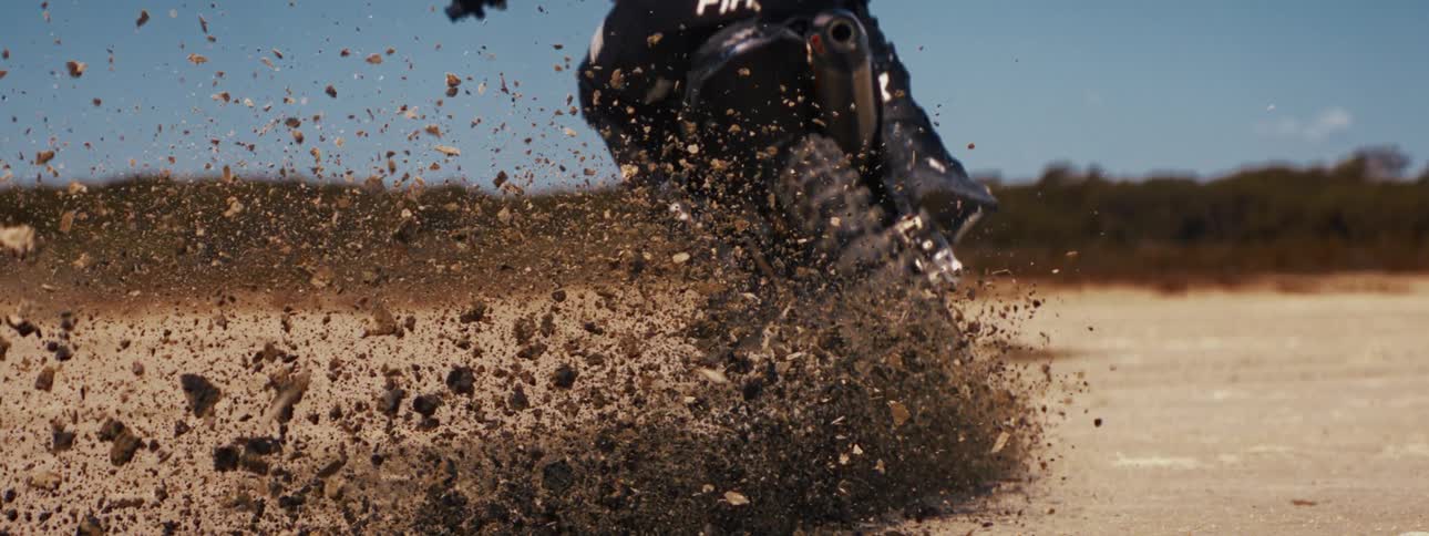 In an epic slow-motion close-up, a motocross bike's rear tire digs into the dirt, launching a shower of rocks and soil into the air
