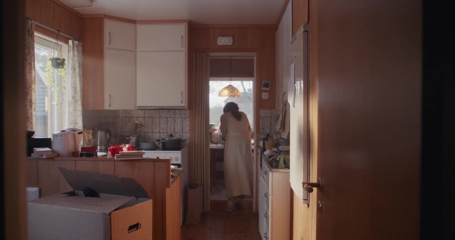 A woman stands with her back to the camera in a sunlit, retro-style kitchen while her child sits at a table in the background