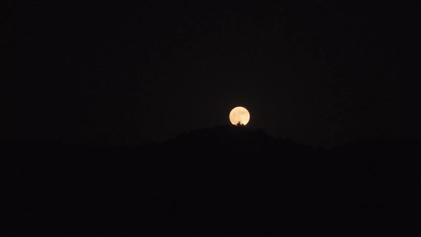 A stunning time-lapse captures the golden full moon rising from behind a silhouetted mountain range against the pitch-black night sky