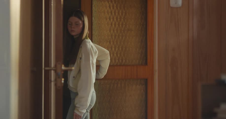 A young girl cautiously enters a room with wood-paneled walls, looking around before slowly closing the door behind her