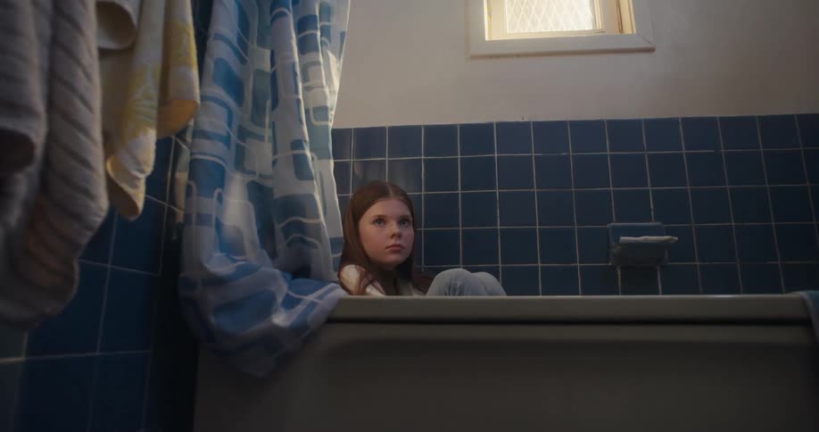 A pensive young girl sits fully clothed in a bathtub, hiding in a dark bathroom