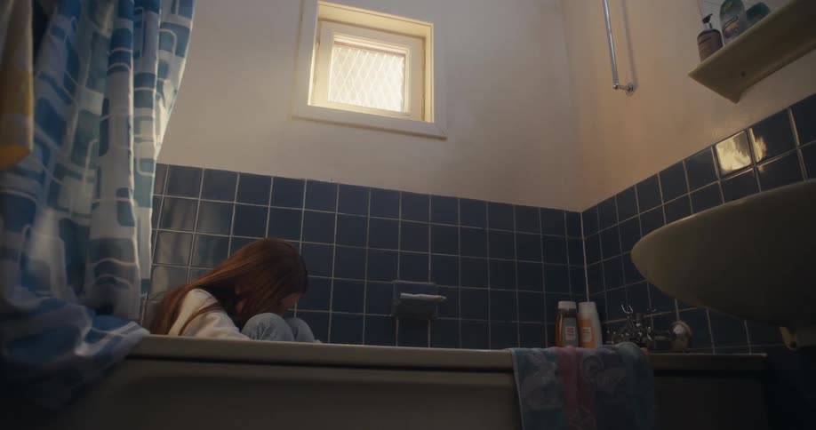 A low-angle shot of a lonely young girl with long red hair sitting fully clothed in a dim, empty bathtub