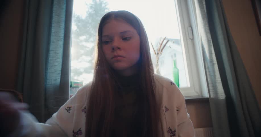 A low-angle, super slow-motion shot of a young girl with long hair eating breakfast and drinking water in front of a sunny window