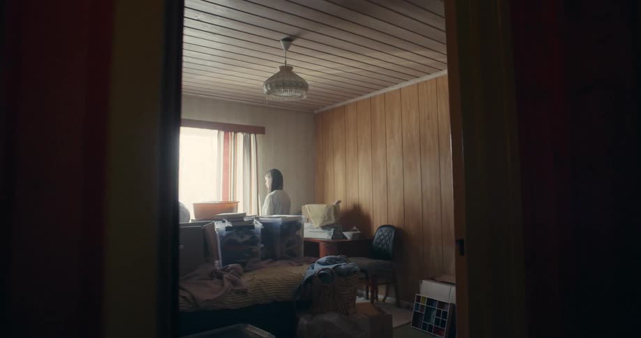 A solitary woman enters a messy, wood-paneled room from her past, looks around, and sits down thoughtfully on the bed